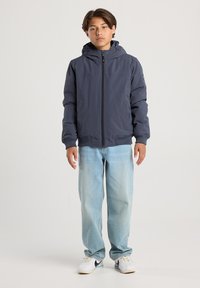 Boy standing facing forward wearing a dark blue hooded jacket, light blue jeans, and white sneakers with black swooshes.