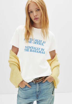T-shirt blanc à manches courtes avec un texte bleu qui dit "PHYSIQUEMENT AU BUREAU, MENTALLEMENT AUX BAHAMAS." Associé à un pantalon en jean délavé et un pull jaune.