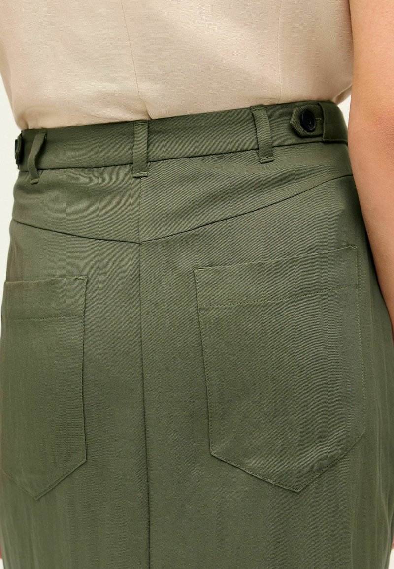 Jupe vert olive avec taille ajustée, deux poches arrière et texture lisse. Le tissu présente de subtiles lignes verticales et un design classique.