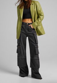 Zelený umělý kožený blejzr přes černý crop top, doplněný volnými černými cargo kalhotami s velkými kapsami a knoflíkovými detaily.