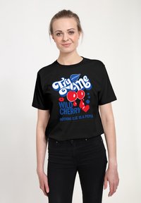 Kvinde iført en sort T-shirt med røde kirsebær og teksten "Try Me Wild Cherry Nothing Else Is A Pepsi" foran, stående mod en lys baggrund.