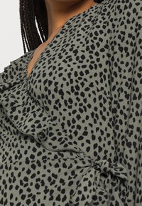 Gros plan sur une personne portant une robe drapée verte avec des taches noires irrégulières et un col froncé.