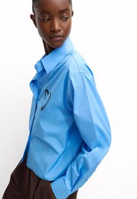 Jasnoniebieska, bawełniana koszula z długim rękawem z button-up z przodu. Posiada wzór serca na piersi i dopasowany krój.