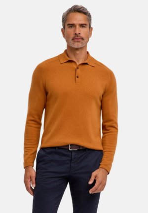Mężczyzna z siwymi włosami, ubrany w rdzawy longsleeve polo i ciemne spodnie, stojący na jednolitym tle.