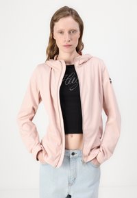 Ροζ ζακέτα με φερμουάρ και κουκούλα, με λεία υφή και μακριά μανίκια, φορεμένη πάνω από μια μαύρη κοντομάνικη μπλούζα. Ορατά γαλάζια ψηλόμεσα τζιν.