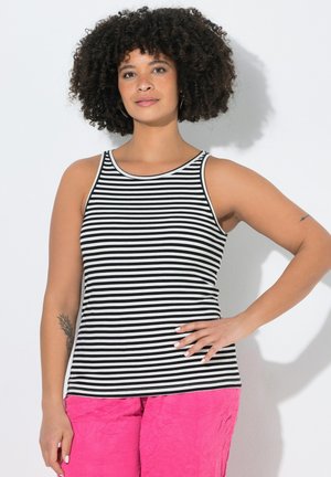 Donna con capelli ricci indossa un top senza maniche a righe bianco e nero e pantaloni rosa acceso, in piedi con una mano sul fianco.