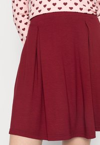 Bordeauxrode geplooide rok van zacht materiaal, met een gladde textuur en een uitlopende vorm. Zoom afgewerkt met een gestikte rand.