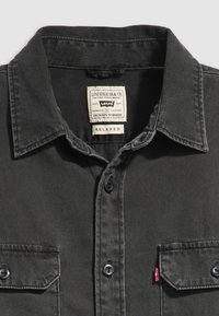 Čierna rifľová košeľa s golierom na gombíky, dvoma náprsnými vreckami a štítkom Levi's. Má tmavé gombíky a voľný strih.