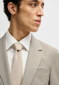 Giacca di abito grigia chiara con una texture sottile, abbinata a una camicia bianca e una cravatta beige glitterata. Il fazzoletto da taschino presenta un motivo a pois.