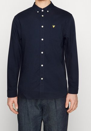 Homme portant une chemise bleu marine à manches longues avec boutons et une poche poitrine ornée d'un petit logo jaune brodé.