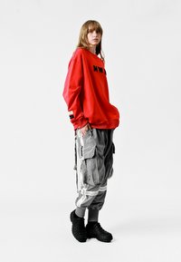 Sudadera roja de gran tamaño con letras negras, combinada con pantalones cargo grises que cuentan con bolsillos con cremallera y detalles de cinta. Zuecos negros en los pies.