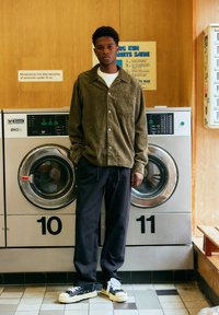 Jeune homme portant une chemise en velours côtelé verte se tient devant des machines à laver numérotées 10 et 11 dans une laverie aux murs en bois.