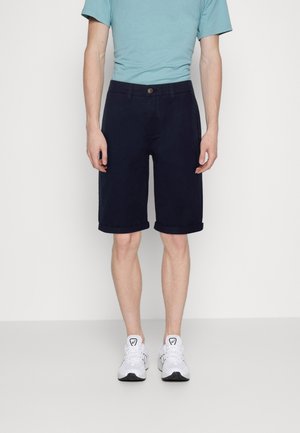 Mann trägt marineblaue Shorts mit umgekrempeltem Saum, hellblaues Kurzarmhemd und weiße Sportschuhe, steht vor einem einfarbigen Hintergrund.