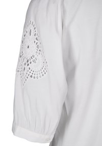 Vit blus med puffärm, prydd med intrikat broderad spets vid armbågen. Mjuk textur och detaljer med knappar vid manschetten.