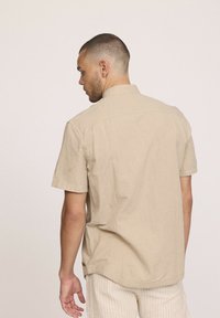 Chemise en lin beige à manches courtes avec col, présentant un ourlet droit et une texture subtile. La vue de dos met en avant le design épuré et les accents minimaux.