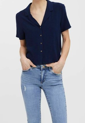 Person wearing a navy short-sleeve buttoned shirt tucked slightly into light blue high-waisted jeans with hands in pockets.