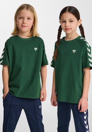 Grønne bomulds t-shirts med korte ærmer, der har et lille logo og kontrasterende hvide chevronstriber på skuldrene. Mørkeblå bukser.