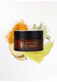 Bernsteinfarbener Glasbehälter mit schwarzem Deckel, beschriftet mit "True Botanicals Ginger Turmeric Cleansing Balm." Umgebende Zutaten sind Kurkumapulver und Ingwer.