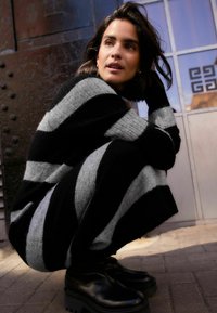 Schwarz-gestreiftes und graues Strickkleid mit geripptem Muster, getragen mit schwarzen Plateauschuhen. Der Hintergrund zeigt eine strukturierte Wand und gemustertes Glas.
