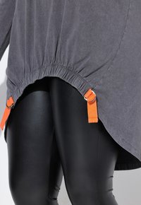 Top grigio, con vestibilità rilassata e orlo arricciato, con accenti in tessuto arancione e hardware argentato, indossato sopra leggings neri lucidi. Tessuto liscio, design informale.