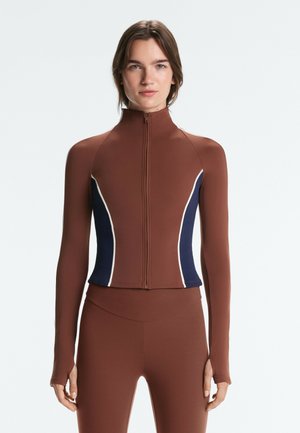 Donna che indossa un top marrone aderente con zip e maniche lunghe, con pannelli laterali blu navy e bianchi e leggings marroni coordinati, in piedi davanti a uno sfondo semplice.