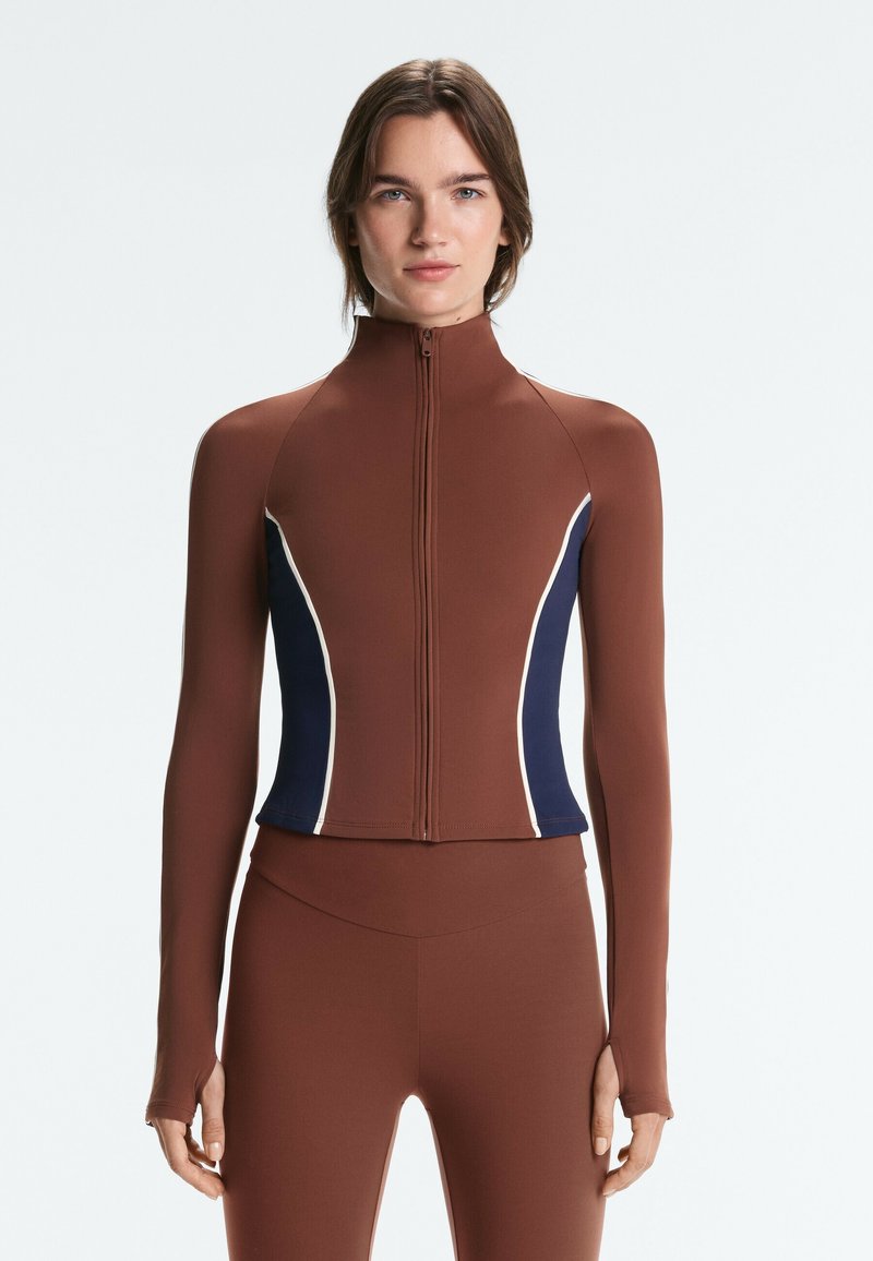 Femme portant un haut zippé marron ajusté à manches longues avec des panneaux latéraux bleu marine et blancs, et un legging marron assorti, debout devant un fond uni.