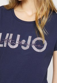 Kvinna iklädd en marinblå t-shirt med ett randigt och prickigt "Blå" textdesign som delvis är täckt av det snirkliga ordet "andas."