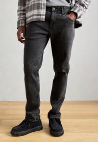 Czarne dżinsy o slim fit, z przednimi kieszeniami, noszone z czarnymi zamszowymi butami na teksturowanej gumowej podeszwie.