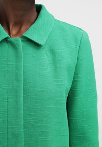 Zelený texturovaný blazer s límcem, strukturovaným designem a vertikálními švy. Látka vypadá jako tkaná s jemným vzorem.