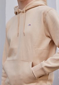 Sudadera con capucha beige hecha de tela suave, que cuenta con un bolsillo tipo canguro, capucha con cordón y un pequeño logo bordado en el pecho.