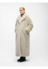 Faux pelsjakke i lys beige, lang længde med sjal krave, sidelommer og tekstureret fluffy yderside. Sorte ankelstøvler synlige nedenfor.