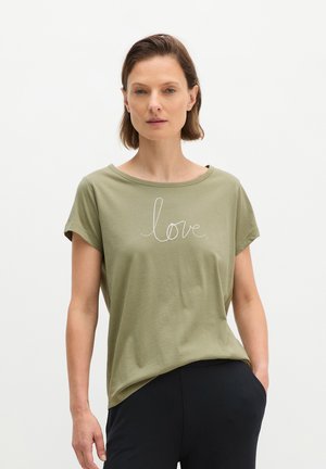 Femme portant un t-shirt vert olive avec le mot "love" en écriture blanche, debout avec une main dans la poche devant un fond uni.