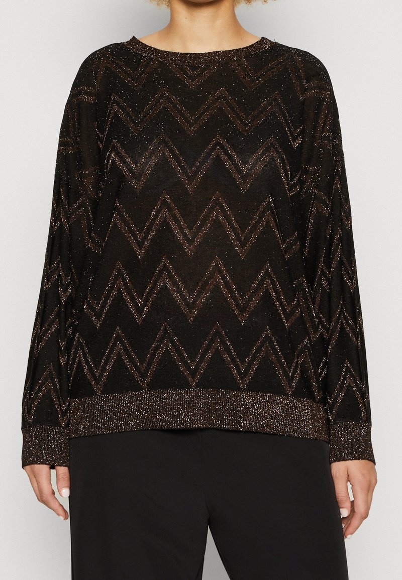 Femme portant un pull noir à manches longues avec des motifs zigzag bronze scintillants et des poignets et un ourlet assortis.