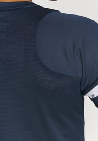 Merinavaate sinisenä, jossa on teksturoitu verkko-takakappale ja sileä kangas hartioilla, valkoisilla yksityiskohdilla hihoissa.