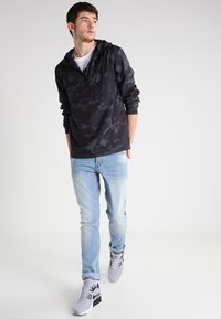 Jachetă pulover neagră cu camuflaj, cu fermoar frontal, asortată cu blugi denim ușor albaștri și adidași gri. Material fin și croială lejeră.