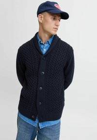 Marine gebreide cardigan met sjawlcol, met een gekruist kabelpatroon en vijf donkere knopen. Gedragen over een denimshirt.