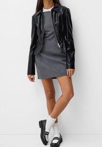 Czarna kurtka z sztucznej skóry o dopasowanym fasonie, noszona na szarym sukienkę bez rękawów. W komplecie z białymi skarpetkami w prążki i czarnymi butami na pasek.