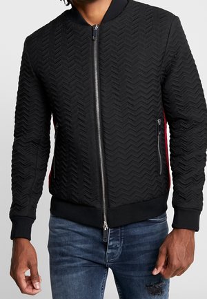 Cazadora bomber negra acolchada con patrón en forma de chevron, cremallera frontal plateada, bolsillos laterales con cremallera, usada con vaqueros azules desgastados.