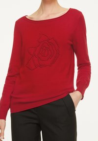 Pull en tricot rouge avec un col large, présentant un motif de rose texturé. Les manches longues et le corps ajusté s’accordent avec un pantalon noir.