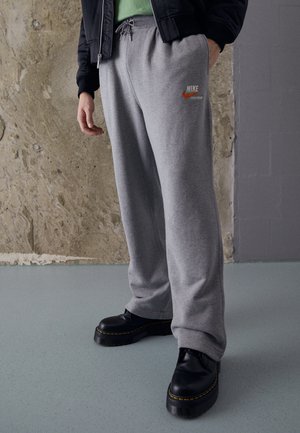 Person iført grå Nike joggingbukser, sorte chunky sko, en grøn top og en sort jakke, stående indendørs mod teksturerede vægge.