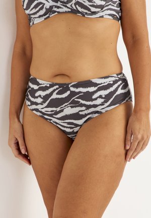 Kvinde iført sort-hvid zebra-mønstret bikini med højtaljede trusser, stående mod en ensfarvet lys baggrund.