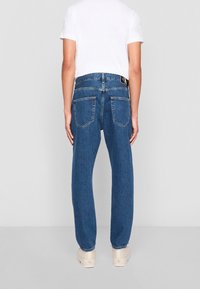 Blå denimjeans med en avslappnad passform, med två bakfickor, en svart etikett i midjan och lätt slitna fållar. Vit t-shirt som bärs ovanpå.