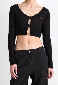 Cárdigan negro corto con mangas largas, hecho de un tejido de punto texturizado. Presenta un escote en V profundo y dos cierres de botón en la parte delantera.