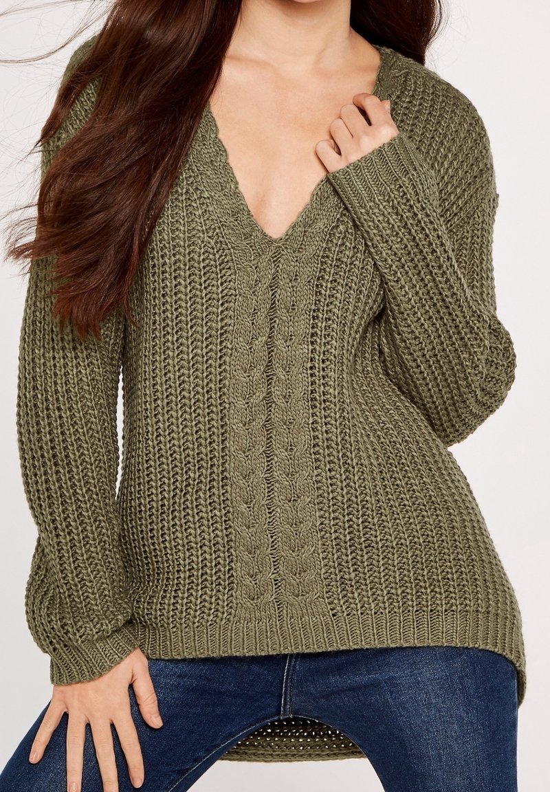 Femme portant un pull en maille épaisse vert olive avec col en V et manches longues, associé à un jean bleu foncé, main près du col.