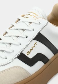 Baskets blanches avec des accents noirs et un bout en daim beige. Présentent un logo noir texturé et des lacets assortis. Semelle extérieure en caoutchouc pour plus de durabilité.