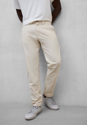 Persona vestida con pantalones casuales beige claro, zapatillas blancas y una camisa blanca, de pie con las manos detrás de la espalda contra una pared gris.