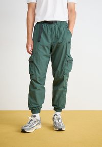 Green nylon cargo pants with elastic cuffs, multiple side pockets, and an adjustable belt. Paired with gray and white athletic shoes.