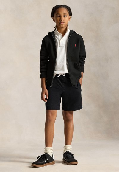 Boy standing with hands in pockets wearing black zip-up hoodie, white polo shirt, black shorts, white socks, and black sneakers.