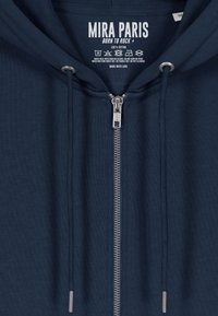 Felpa con zip in cotone blu navy, con cerniera argento, cordini e etichetta interna che mostra il marchio e le istruzioni per la cura.