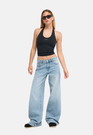Jeune femme portant un top noir dos-nu, un jean bleu clair à jambes larges, des chaussures noires et des lunettes de soleil foncées, debout devant un fond blanc.